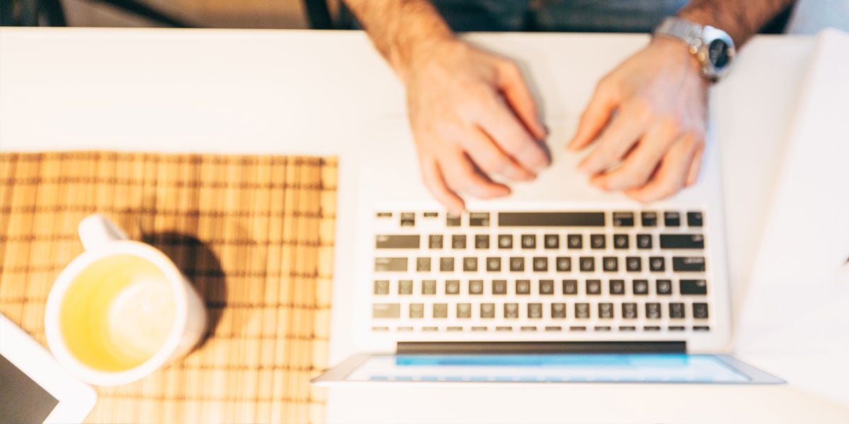 Males hands hovering over a laptops keyboard with a cup of coffee to the side